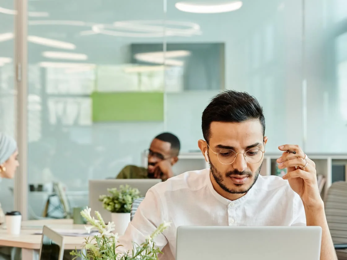 Homem de óculos concentrado no notebook, em escritório claro com luz natural.